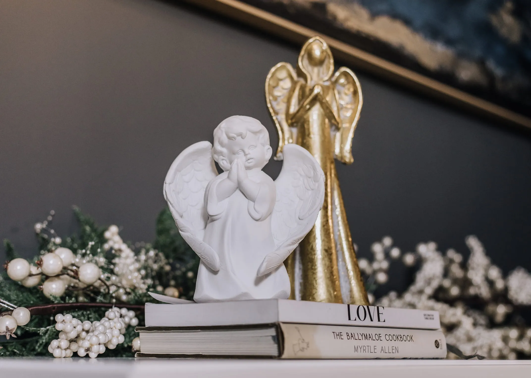 A lifestyle image of the Praying angel with a gold angel on a mantle with pearl garland.