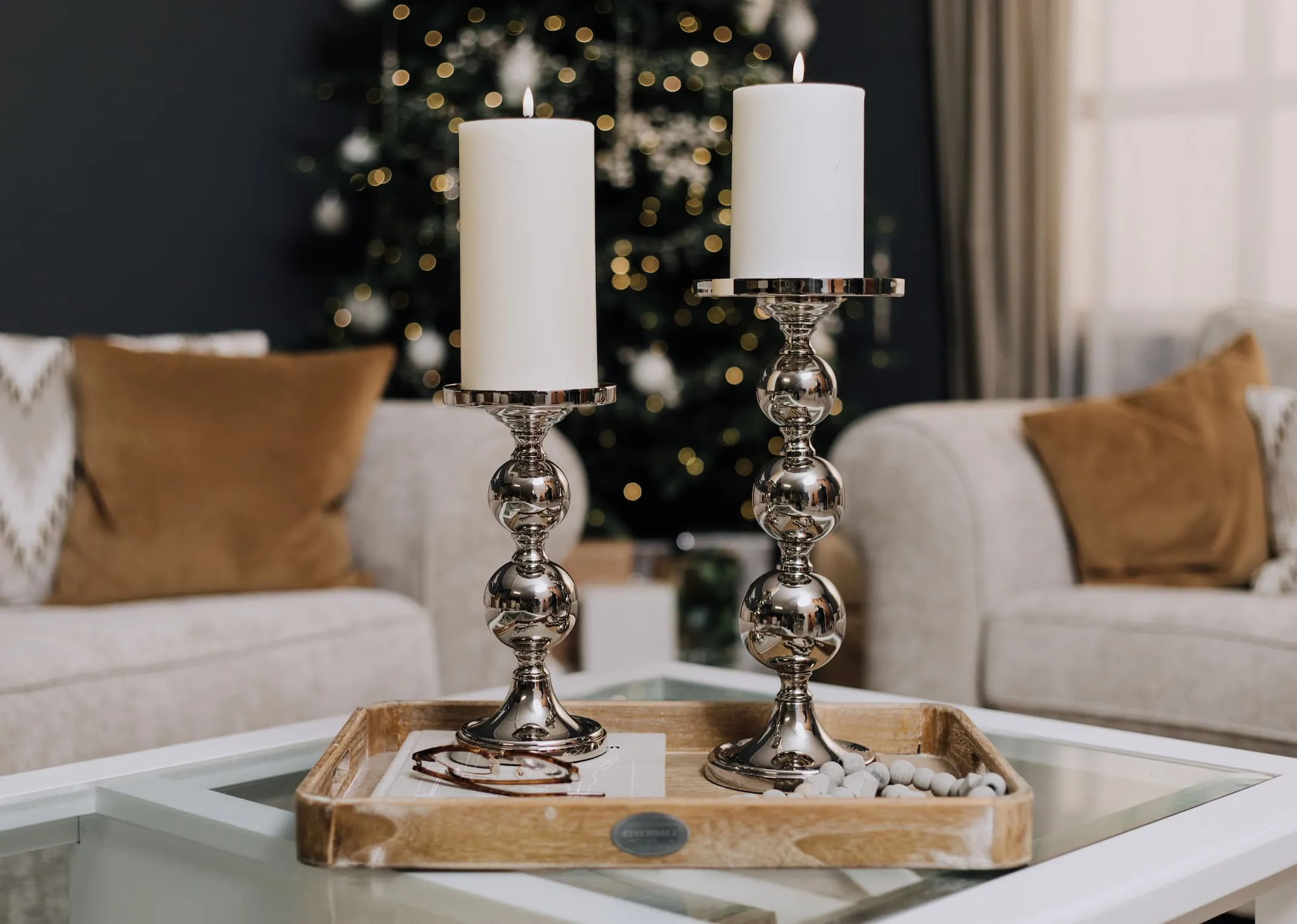 A lifestyle image of a wood tray on a coffee table with 2 silver pillar candle holder stands with candles.