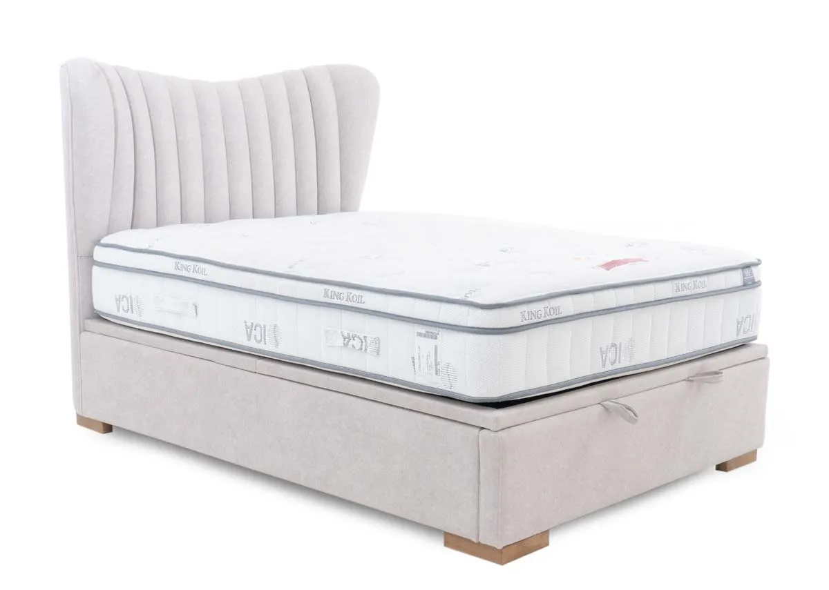 A light grey fabric ottoman bed with a white mattress on top presented from an angle. The headboard features elegant vertical tufting.