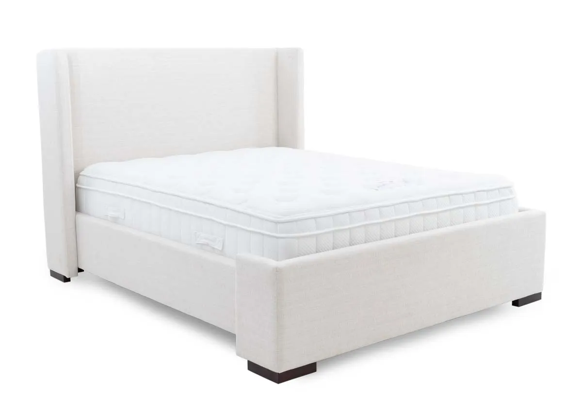 A beige fabric bed frame with high winged headboard from EZ Livings Quay west range. Angled view of matttress inserted not included.