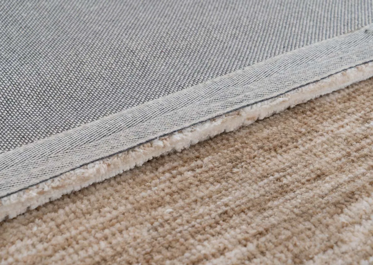 A close up shot showing the sandy beige rugs woven top surface and its grey textured underside with a visible stitched edge.