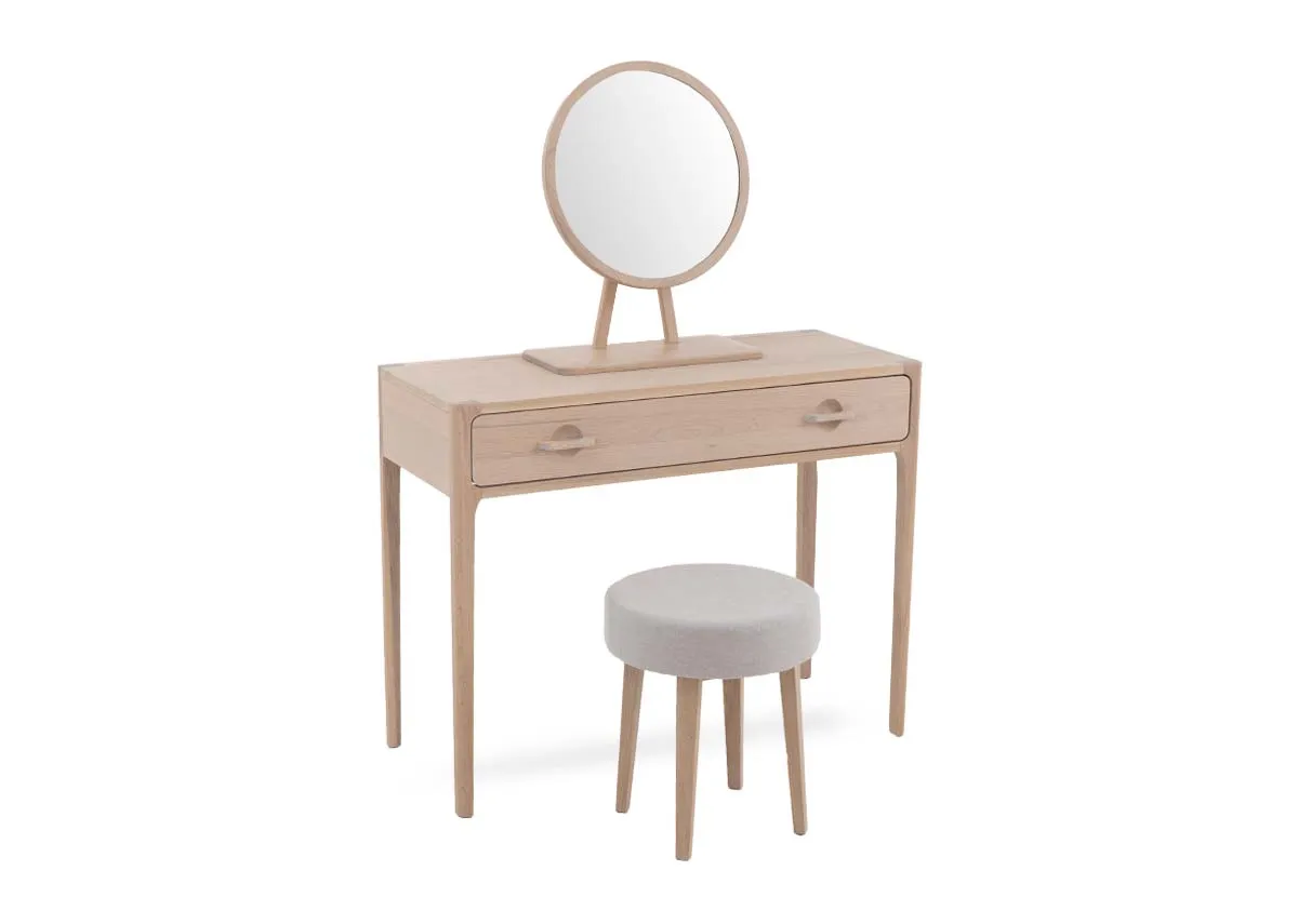 A white oak dressing table with 1 large drawer and round mirror with a grey fabric stool. Angled view