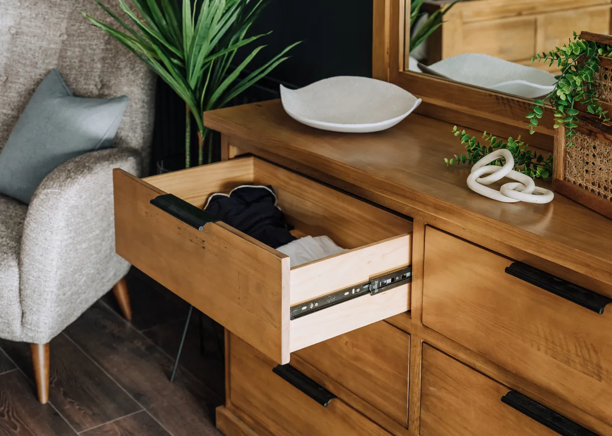 A close up of an open drawer in a light brown wooden tallboy showing the wooden interior and black handle.