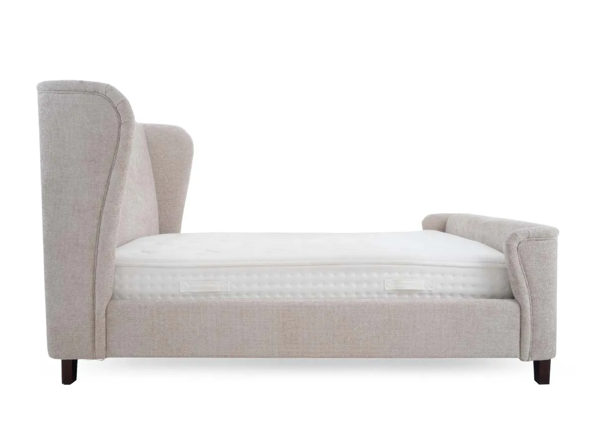 A beige fabric bed frame with a high winged headboard and a low footboard shown from a side profile with a white mattress.