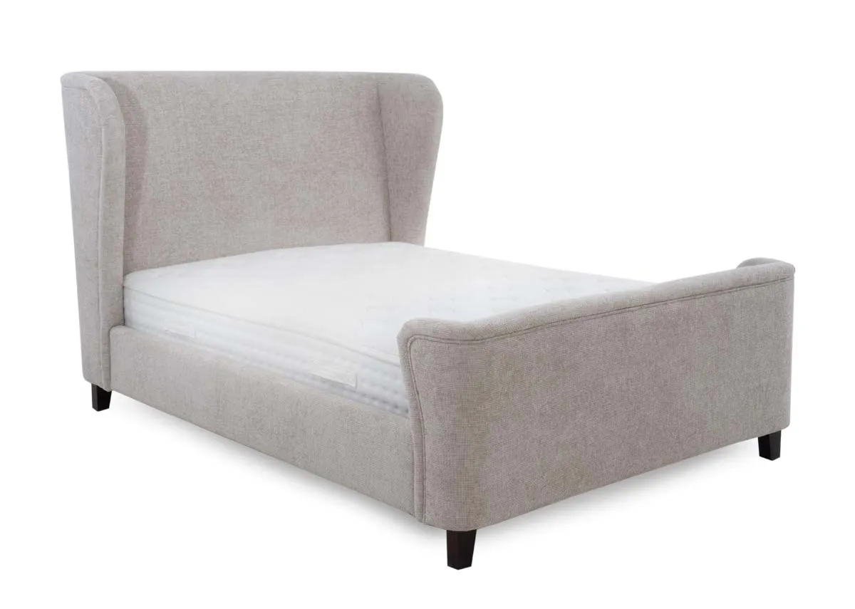 A beige fabric bedframe with winged headboard and dark brown legs. Angled view of bed with mattress.