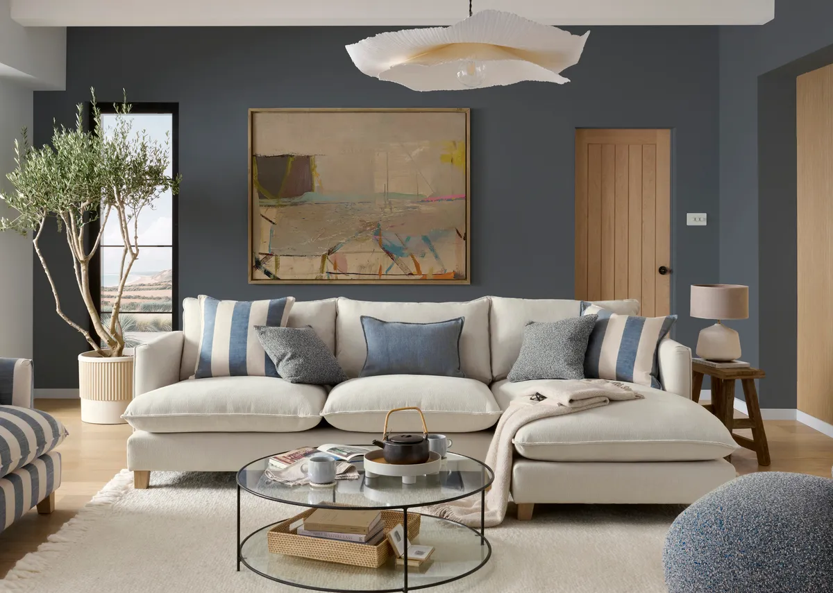 A lifestyle image of the Howth linen rhf corner chaise with blue and stripe cuddler with round glass coffee table.