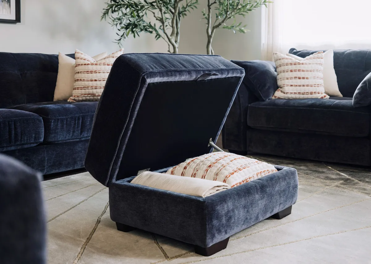 A lifetsyle image of the Darcy denim blue footstool open showing hidden storage fitting a cushion and blanket.
